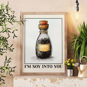 May include: A framed print showing a detailed illustration of a soy sauce bottle with a cork stopper. The bottle is dark with a yellow label. The text "I'M SOY INTO YOU" is printed below the illustration. The print is in a wooden frame.