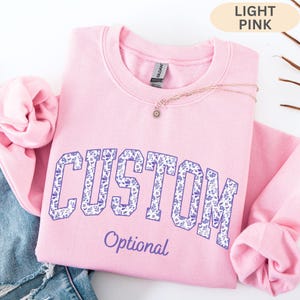 May include: A light pink crewneck sweatshirt featuring the word CUSTOM in large block letters filled with a purple floral pattern. Below the word CUSTOM, the word Optional is printed in a purple script font. A gold chain necklace rests on top.