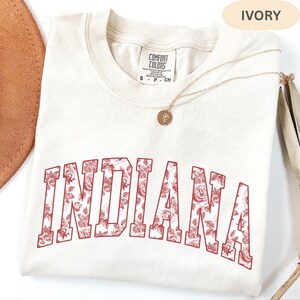 以下が含まれることがあります： 赤色の花柄で「INDIANA」の文字が入ったアイボリー色のスウェットシャツ。クルーネックと半袖です。小さなペンダントが付いたゴールドのネックレスがシャツの上にあります。「Comfort Colors」と書かれたラベルが付いています。