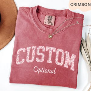 May include: Crimson-colored t-shirt with the word "CUSTOM" in white floral print lettering. The word "Optional" is printed below in a script font. A silver necklace with a pendant is on the shirt.