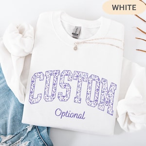 May include: A white crewneck sweatshirt featuring the word CUSTOM in large block letters filled with a purple floral pattern, with the word Optional printed below in purple script. A delicate gold chain necklace rests on the collar area.