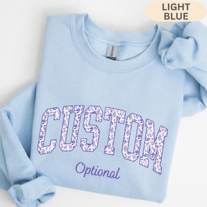 May include: A light blue crewneck sweatshirt featuring the word CUSTOM in large, block-style letters filled with a purple floral pattern. Below the main text, the word Optional is printed in a smaller, purple script font. A matching scrunchie is included.