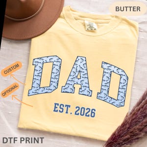以下が含まれることがあります： バター色のTシャツ。水色のブロック体で「DAD」の文字が描かれ、イルカの模様で埋められています。「EST. 2026」の文字が「DAD」の下にあります。TシャツのサイズはXL-TG-EGです。