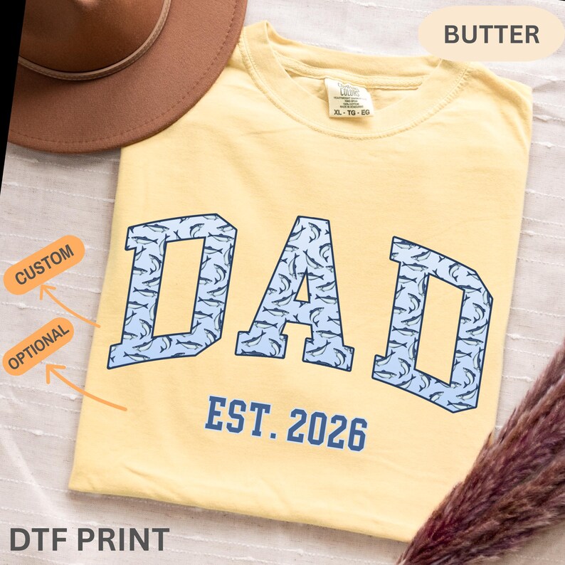 以下が含まれることがあります： バター色のTシャツ。水色のブロック体で「DAD」の文字が描かれ、イルカの模様で埋められています。「EST. 2026」の文字が「DAD」の下にあります。TシャツのサイズはXL-TG-EGです。