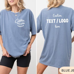 May include: A blue jean coloured t-shirt with the text "Custom TEXT / LOGO here" printed on the front and back. The front of the shirt has a curved text design. The shirt is a short-sleeved, oversized fit.