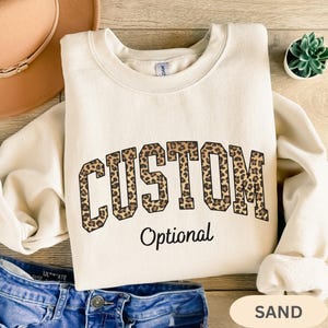 May include: Sand-colored sweatshirt with the word "CUSTOM" in a leopard print design, arched across the chest. Below, the word "Optional" is printed in a black, simple font. The sweatshirt is folded on a wooden surface.