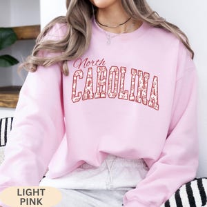 May include: A light pink long-sleeve crewneck sweatshirt featuring the text North CAROLINA in a bold, arched font. The letters are outlined in red and filled with a white and red floral pattern. The sweatshirt has a ribbed collar and cuffs.