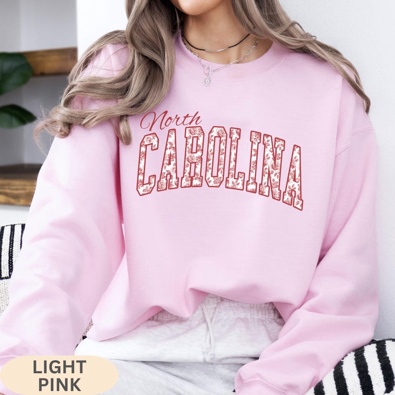 May include: A light pink long-sleeve crewneck sweatshirt featuring the text North CAROLINA in a bold, arched font. The letters are outlined in red and filled with a white and red floral pattern. The sweatshirt has a ribbed collar and cuffs.
