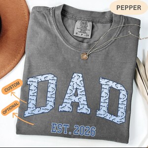 以下が含まれることがあります： グレーのComfort Colors Tシャツ。 鮫の模様で埋められた、明るい青色のブロック文字で「DAD」という文字がプリントされています。「EST. 2026」の文字が「DAD」の下にあります。 ペンダント付きのシルバーネックレスがシャツの上にあります。