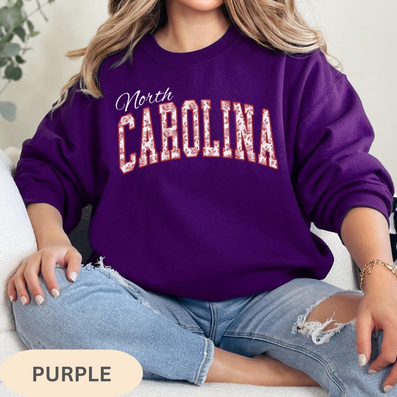 May include: A deep purple crewneck sweatshirt featuring the text North CAROLINA in a white, arched, block-letter font. The letters are filled with a detailed white floral pattern. The sweatshirt has long sleeves and a ribbed collar and cuffs.