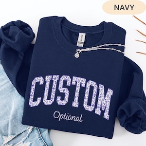 May include: A navy blue crewneck sweatshirt featuring the word CUSTOM in large block letters filled with a purple and white floral pattern. Below the word CUSTOM, the word Optional is printed in a white script font. A silver chain necklace is visible.