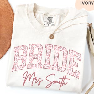 May include: Ivory-colored t-shirt with the word "BRIDE" in large, pink floral letters. Below, in a cursive font, is "Mrs Smith". A gold necklace with a pendant rests on the shirt. The shirt is a Comfort Colors brand.