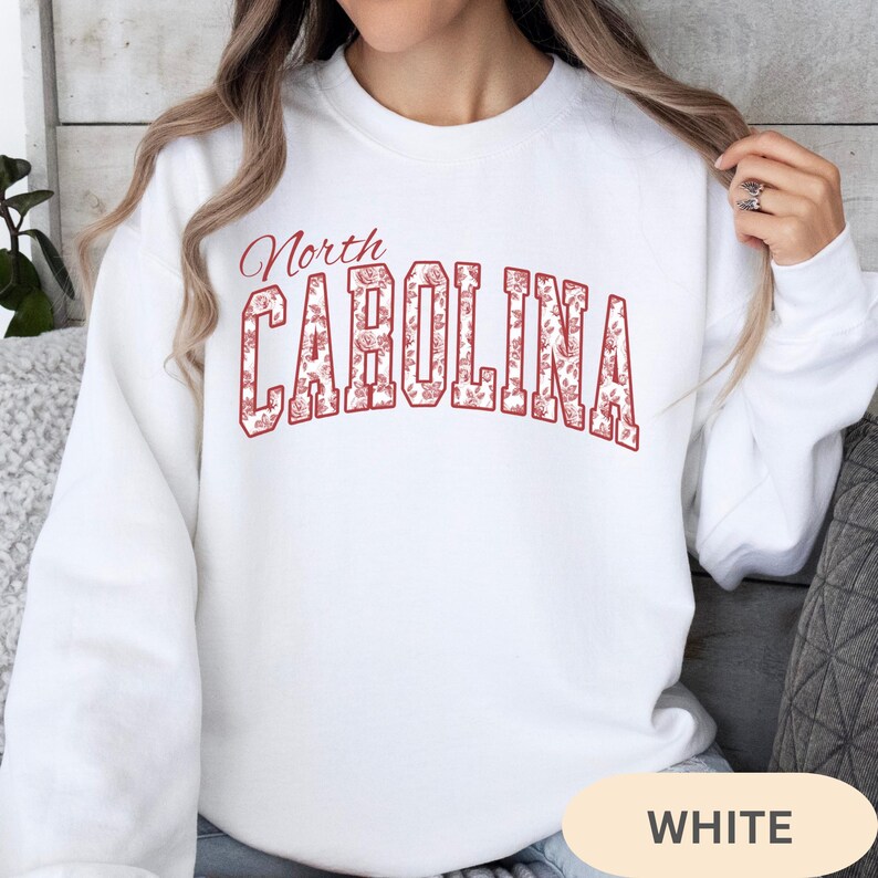 May include: A white crewneck sweatshirt featuring the text North CAROLINA in a red outline font. The letters are filled with a detailed red and white floral pattern. The sweatshirt has long sleeves and a ribbed collar, cuffs, and hem.