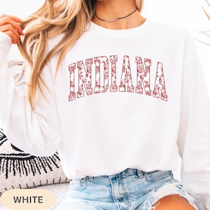 以下が含まれることがあります： 胸にマルーンの花柄で「INDIANA」の文字が入った白い長袖スウェットシャツ。スウェットシャツは無地の白です。画像の下部には「WHITE」の文字がプリントされています。