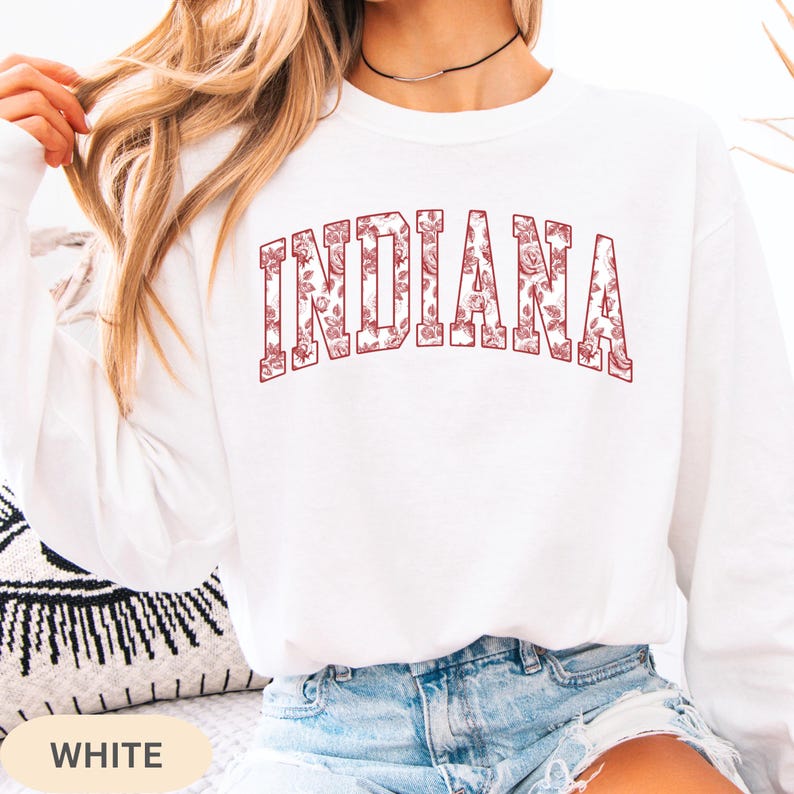 以下が含まれることがあります： 胸にマルーンの花柄で「INDIANA」の文字が入った白い長袖スウェットシャツ。スウェットシャツは無地の白です。画像の下部には「WHITE」の文字がプリントされています。
