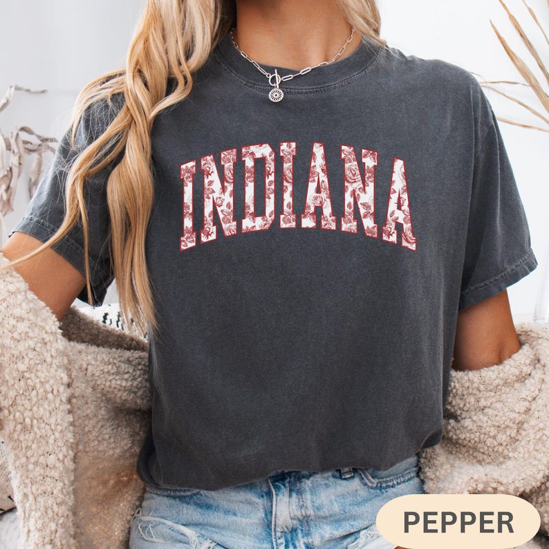 以下が含まれることがあります： 胸に花柄のデザインで「INDIANA」の文字が入った濃いグレーのTシャツ。テキストは赤と白の花柄です。Tシャツは半袖とクルーネックです。