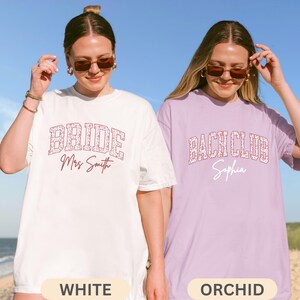 May include: Two oversized t-shirts, one white and one orchid, with text. The white shirt says "BRIDE Mrs Smith" in a floral design. The orchid shirt says "BACH CLUB Sophia" in a similar floral design. The shirts are for a bridal party.
