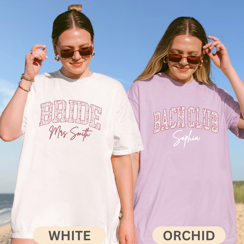 May include: Two oversized t-shirts, one white and one orchid, with text. The white shirt says "BRIDE Mrs Smith" in a floral design. The orchid shirt says "BACH CLUB Sophia" in a similar floral design. The shirts are for a bridal party.