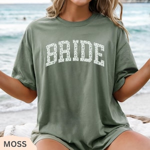 May include: A moss green t-shirt with the word "BRIDE" in white, floral-patterned letters. The shirt has a classic crew neck and short sleeves. The word "MOSS" is printed in a cream-colored rectangle at the bottom left.