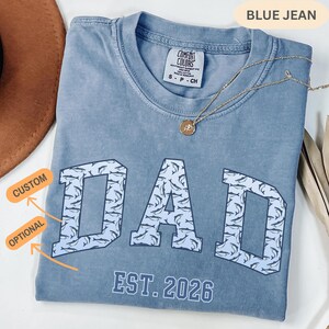 以下が含まれることがあります： 「DAD」の文字が魚の模様で埋め込まれた、カレッジ風フォントのブルーデニムカラーTシャツ。「EST. 2026」の文字が「DAD」の下にあります。ペンダント付きのゴールドネックレスがシャツの上にあります。シャツは折りたたまれています。