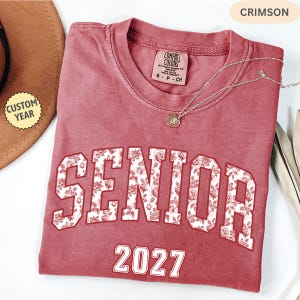 Peut inclure: T-shirt de couleur cramoisie avec le mot "SENIOR" en lettres blanches à motifs floraux et l'année "2027" en blanc. Un collier argenté avec un pendentif repose sur le t-shirt. Le t-shirt est de la marque Comfort Colors.