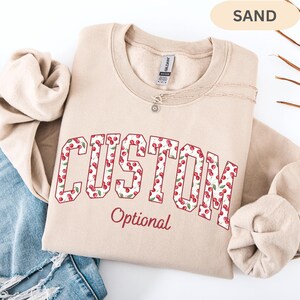 Pode incluir: Uma camisola de gola redonda cor de areia com a palavra CUSTOM em letras grandes de bloco preenchidas com um padr&atilde;o de cerejas vermelhas e folhas verdes. Abaixo da palavra CUSTOM, a palavra Optional est&aacute; impressa numa fonte cursiva vermelha.