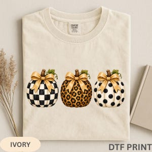 May include: An ivory t-shirt featuring a graphic print of three pumpkins in a row. Each pumpkin is topped with a gold bow and features a different pattern: black and white checkered, brown leopard print, and black polka dots on white.