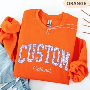 May include: An orange crewneck sweatshirt featuring the word CUSTOM in a purple floral pattern across the chest, with the word Optional printed below in white script. A silver chain necklace with a circular pendant rests on the collar.