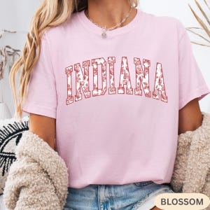 以下が含まれることがあります： 胸に「INDIANA」の文字が花柄でデザインされた、明るいピンク色のTシャツ。テキストは太字でアーチ状になっており、赤いアウトラインと白と赤の花柄が施されています。「BLOSSOM」の文字が下にあります。