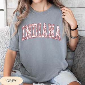 以下が含まれることがあります： 赤色の花柄で「INDIANA」の文字が入ったグレーのTシャツ。クルーネックのスタイルで、柔らかい素材で作られているようです。画像の下部には「GREY」の文字があります。