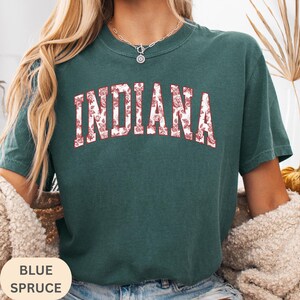 以下が含まれることがあります： 赤色の花柄で「INDIANA」の文字が入った、ブルー・スプルースのTシャツ。クルーネックで半袖です。画像の下部には「BLUE SPRUCE」の文字があります。