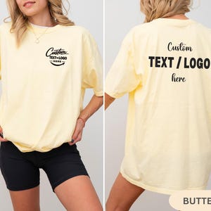 May include: A pale yellow t-shirt with the text "Custom TEXT/LOGO here" in black on the front and back. The shirt has a relaxed fit and short sleeves. The word "BUTTER" is at the bottom of the image.