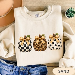 May include: A cream-colored crewneck sweatshirt featuring a graphic of three pumpkins. The first pumpkin has a black and white checkered pattern, the second has a brown leopard print, and the third is white with black polka dots. Each has a gold bow.