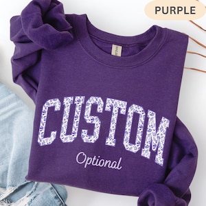 May include: A dark purple long-sleeve sweatshirt featuring the word CUSTOM in large block letters filled with a white and purple floral pattern, with the word Optional written in a smaller white script font underneath the main text.