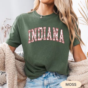 以下が含まれることがあります： 胸に花柄で「INDIANA」の文字が入ったモスグリーンのTシャツ。Tシャツは半袖とクルーネックです。画像右下には「MOSS」の文字が見えます。