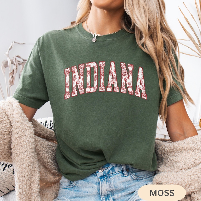 以下が含まれることがあります： 胸に花柄で「INDIANA」の文字が入ったモスグリーンのTシャツ。Tシャツは半袖とクルーネックです。画像右下には「MOSS」の文字が見えます。
