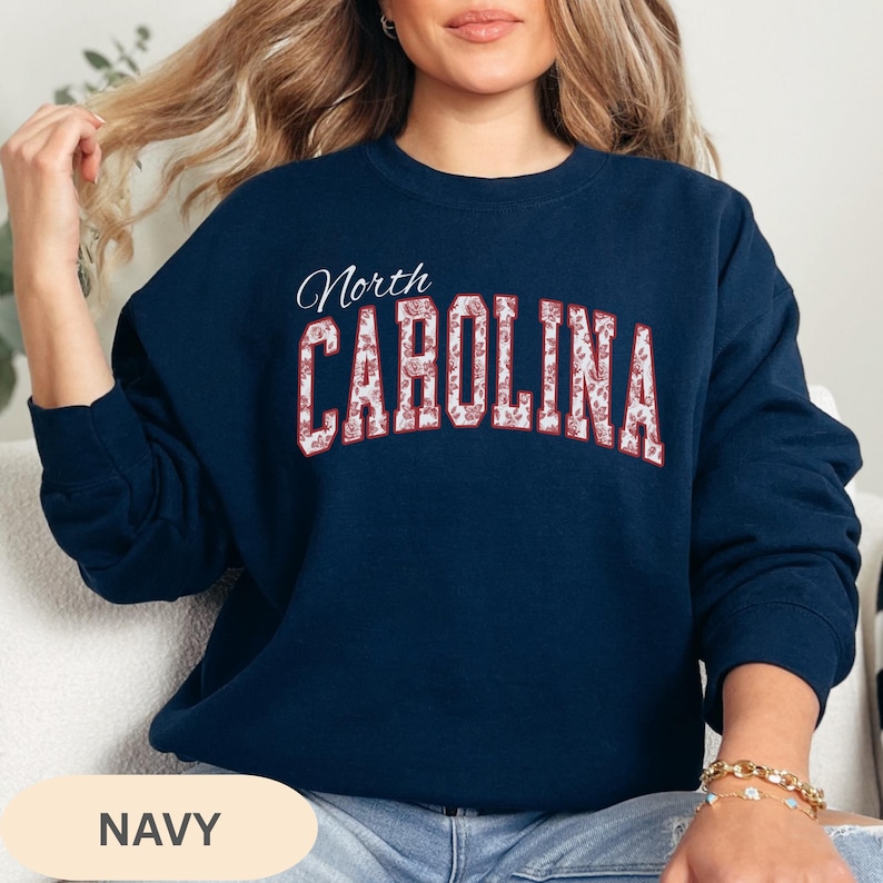 May include: A navy blue crewneck sweatshirt featuring the word North in white script above the word CAROLINA in large, block-style letters. The letters of CAROLINA are filled with a white and red floral pattern on a dark navy background.
