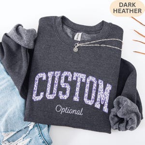 May include: A dark heather gray crewneck sweatshirt featuring the word CUSTOM in large block letters filled with a purple and white floral pattern. Below the word CUSTOM, the word Optional is printed in a smaller, white cursive script font.
