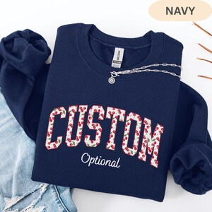 Pode incluir: Uma camisola azul-marinho de gola redonda com a palavra CUSTOM em letras grandes de bloco preenchidas com um padr&atilde;o de cerejas vermelhas e folhas verdes. Abaixo da palavra CUSTOM, a palavra Optional est&aacute; impressa em letra cursiva branca.