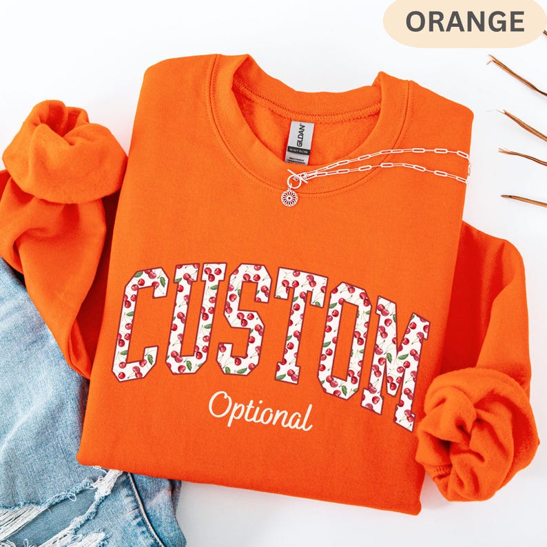 Pode incluir: Uma camisola de gola redonda laranja com a palavra CUSTOM em letras grandes de bloco preenchidas com um padr&atilde;o de cerejas vermelhas. Abaixo da palavra CUSTOM, a palavra Optional est&aacute; impressa em letra cursiva branca. Um colar de corrente prateada repousa no decote.
