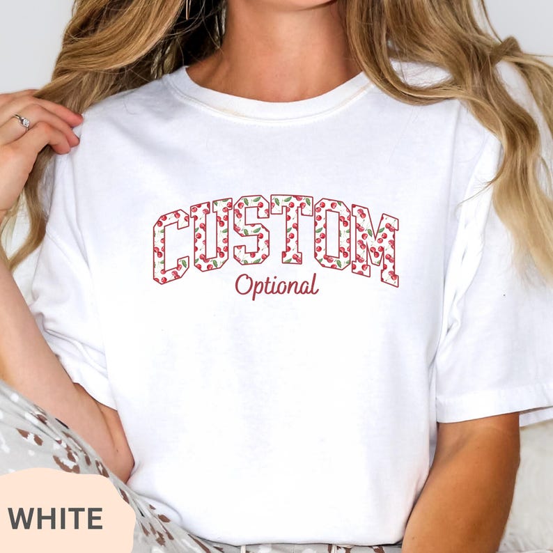 Pu&ograve; includere: Una t-shirt bianca a maniche corte con la parola CUSTOM in un grande carattere in stile blocco riempito con un motivo di ciliegie rosse e foglie verdi. Sotto la parola CUSTOM, la parola Optional &egrave; stampata in un carattere corsivo rosso.