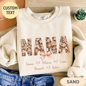 May include: A sand-colored crewneck sweatshirt featuring the word NANA in large block letters filled with a floral pattern in shades of pink, green, and cream. A light pink bow sits below the letters, followed by the names Emma, Olivia, Liam, Daniel, and Katie.