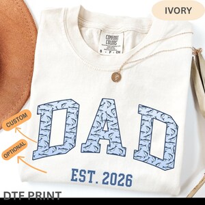 以下が含まれることがあります： イルカの模様で埋められた青いブロック文字で「DAD」という言葉が書かれたアイボリー色のTシャツ。「EST. 2026」という文字が「DAD」の下にあります。ペンダント付きのゴールドネックレスがシャツの上にあります。