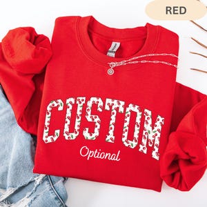 Pode incluir: Uma camisola vermelha de gola redonda com a palavra CUSTOM em letras grandes de bloco preenchidas com um padr&atilde;o de cerejas, com a palavra Optional escrita abaixo em letra cursiva branca. Um colar de corrente prateada repousa no decote.