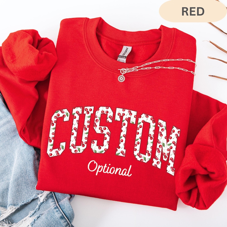 Pode incluir: Uma camisola vermelha de gola redonda com a palavra CUSTOM em letras grandes de bloco preenchidas com um padr&atilde;o de cerejas, com a palavra Optional escrita abaixo em letra cursiva branca. Um colar de corrente prateada repousa no decote.