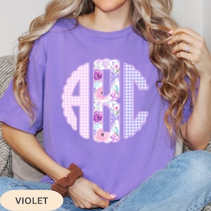 May include: Violet-colored t-shirt with a floral monogram design. The monogram features the letters "ABC" in a decorative, scalloped circle. The letters are filled with a mix of gingham and floral patterns in shades of pink, purple, and white.
