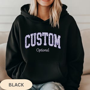 May include: A black hooded sweatshirt featuring the word CUSTOM in a large, arched, light purple floral print font across the chest, with the word Optional written in a smaller, white, cursive script font directly underneath the arched text.