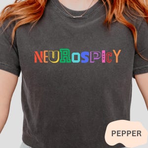May include: A black t-shirt with the text "NEUROSPICY" in rainbow colours. The word "PEPPER" is written in white on a pink circle in the bottom right corner of the image.