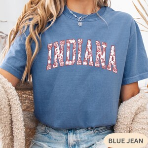 以下が含まれることがあります： 前面に「INDIANA」の文字が花柄でプリントされた青い半袖Tシャツ。テキストは太字で、カレッジ風のフォントで赤いアウトライン。シャツは無地の青色です。