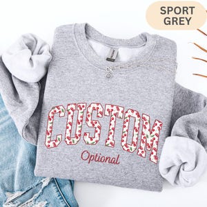 Pode incluir: Uma camisola de gola redonda cinzento desportivo com a palavra CUSTOM em letras grandes de bloco preenchidas com um padr&atilde;o de cerejas vermelhas e folhas verdes. Abaixo da palavra CUSTOM, a palavra Optional est&aacute; impressa numa fonte cursiva vermelha menor.